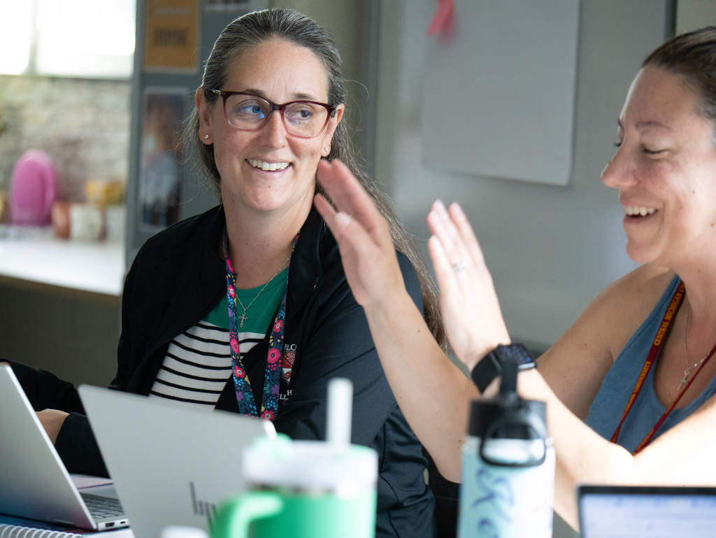 Two women, one with glasses, seated at a table with laptops, clapping hands in a classroom.