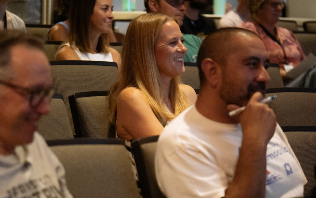 A room with people sitting in chairs. One woman smiles, and a man with a beard holds a pen.