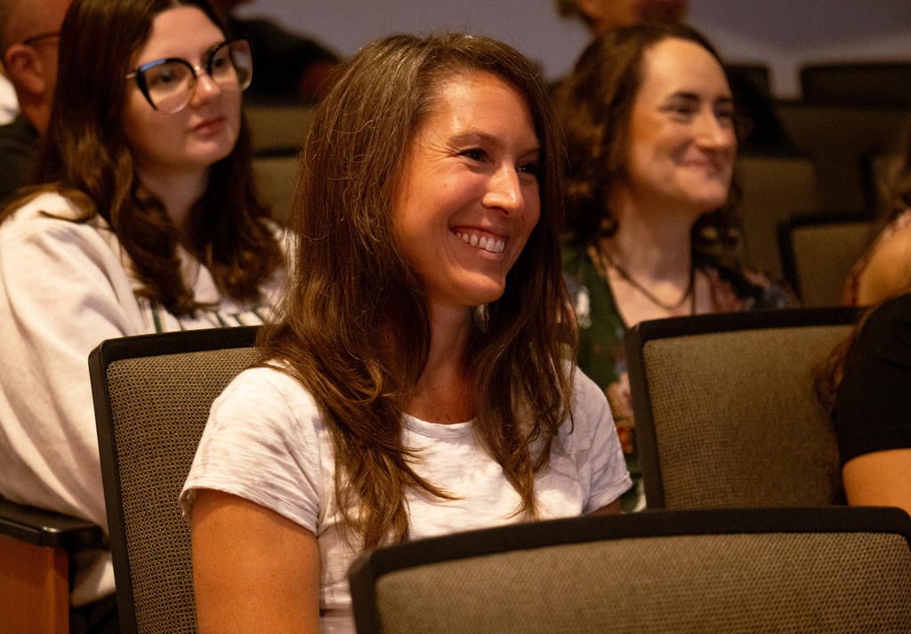 Three women, including one smiling, sit in chairs in a room with blurred background. Two wear white, one wears green.