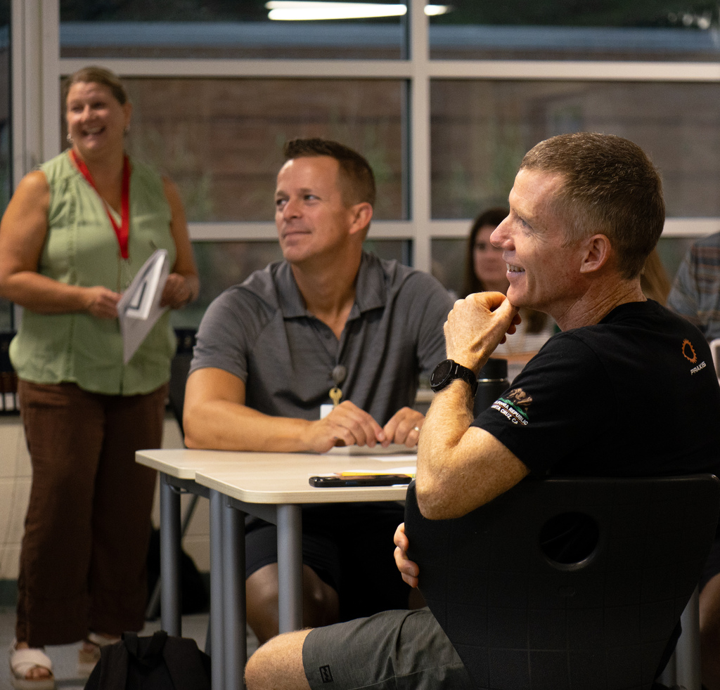 Three adults sit around a table, one smiling. A woman stands behind them holding papers. Glass walls reflect the room.