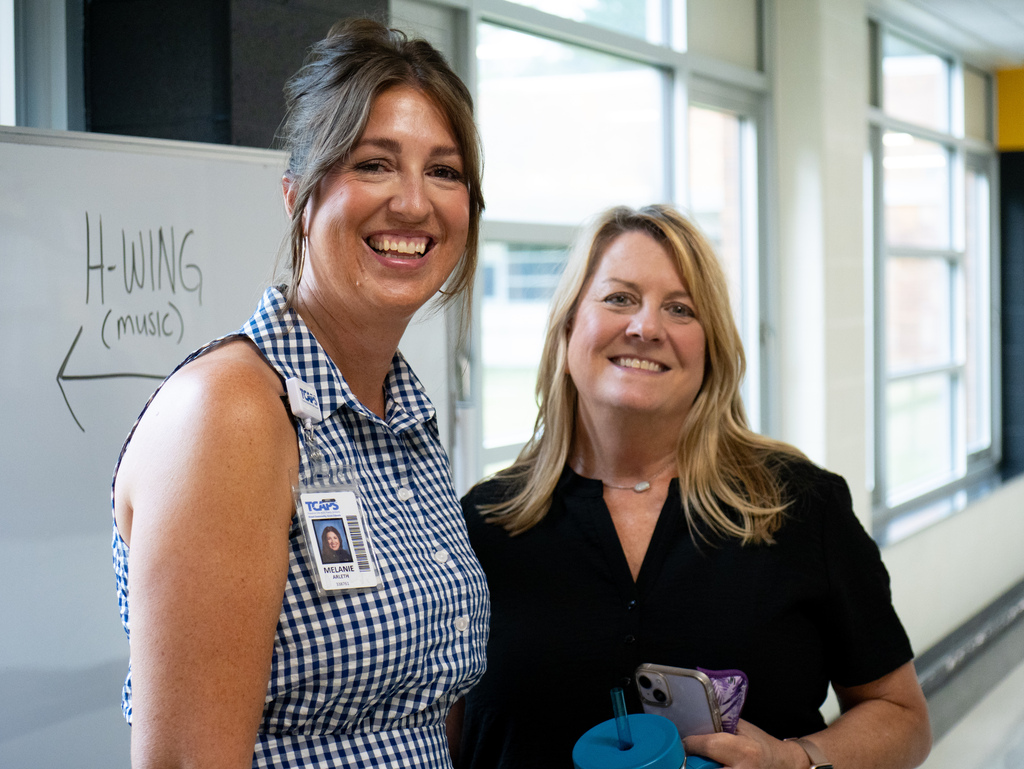 Two women smiling; one holds a blue cup, the other holds a phone. They stand near a white board.
