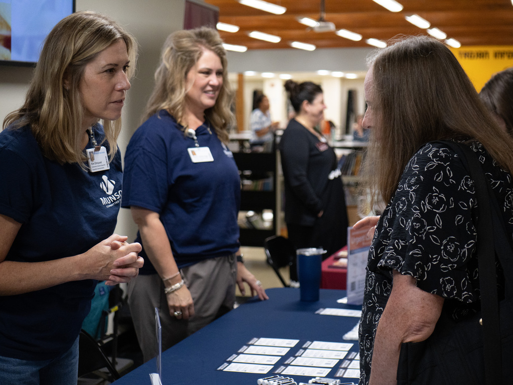 Three women in blue shirts stand near a table with papers and cups. One woman wears a floral blouse.