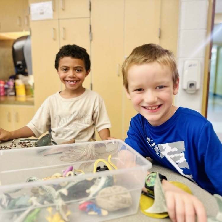 Two children smiling. One has a tray of small toys. Behind them, there is a cabinet and a sink.