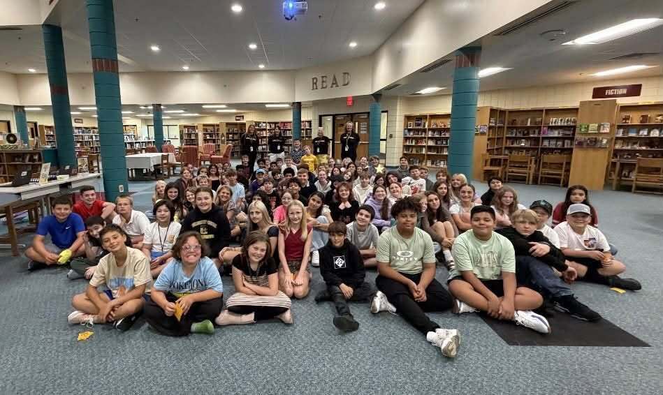 A group of students seated on the floor in a library, some wearing casual clothing, with bookshelves in the background.