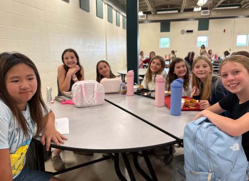 A group of students sitting at tables in a cafeteria. A few bags and lunch boxes are on the tables.