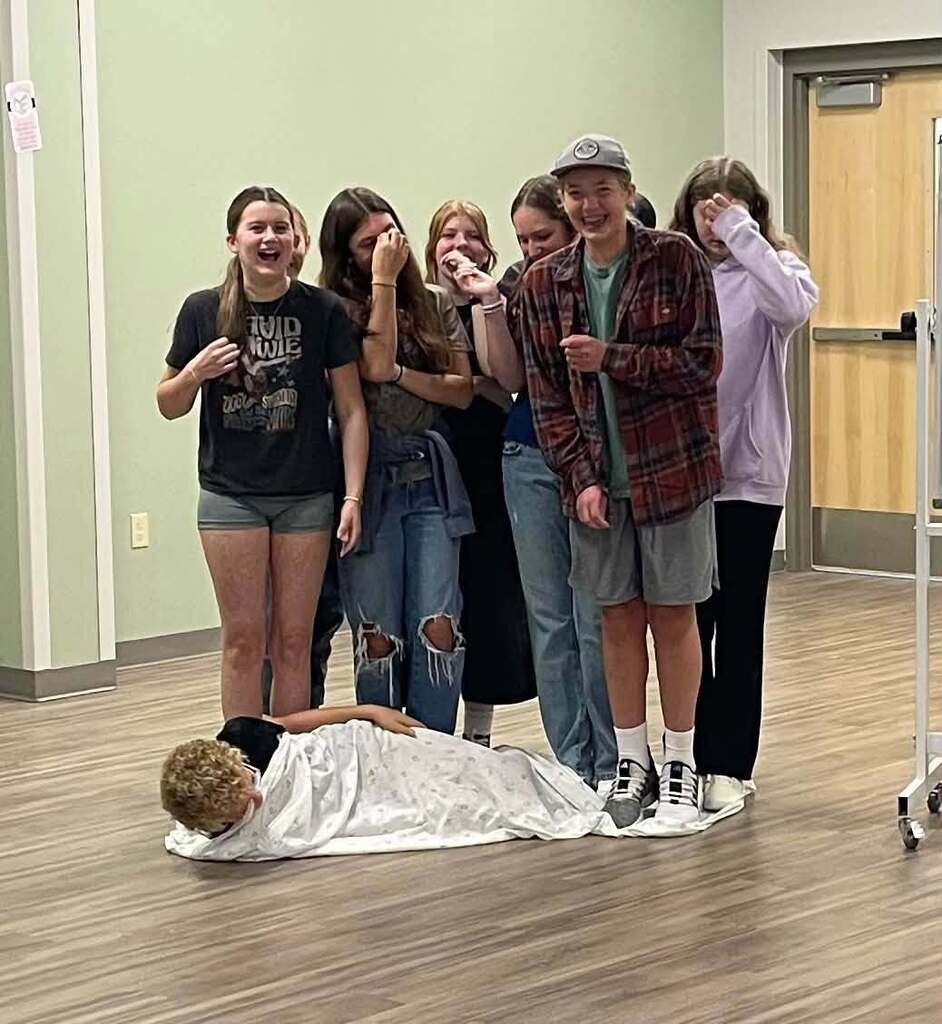 A group of young people pose in a room; one lies on the floor. Green walls, a wooden floor, and a door.
