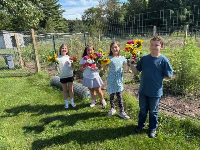 Four kids, three girls and one boy, stand in a garden with flowers in their hands.