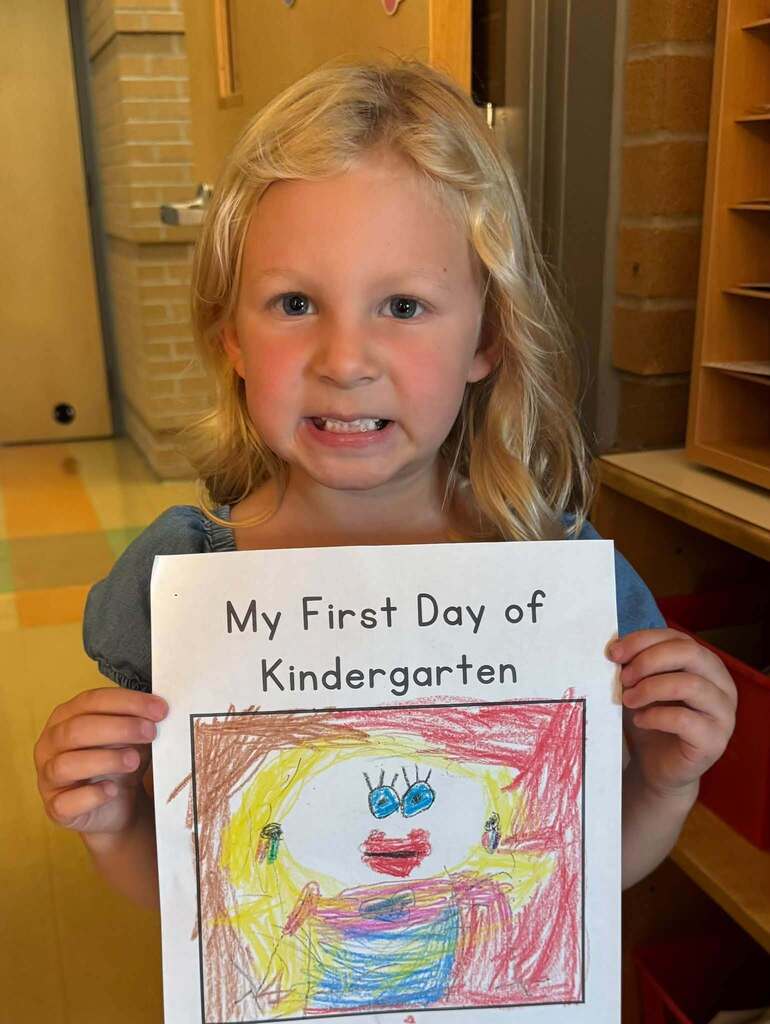 A young girl holds a paper with a drawing and text that says "My First Day of Kindergarten" in a classroom.