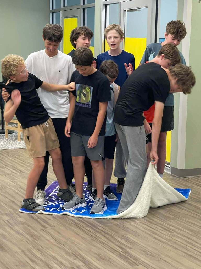 A group of children on a blue mat inside a room, some bending over, others standing behind.