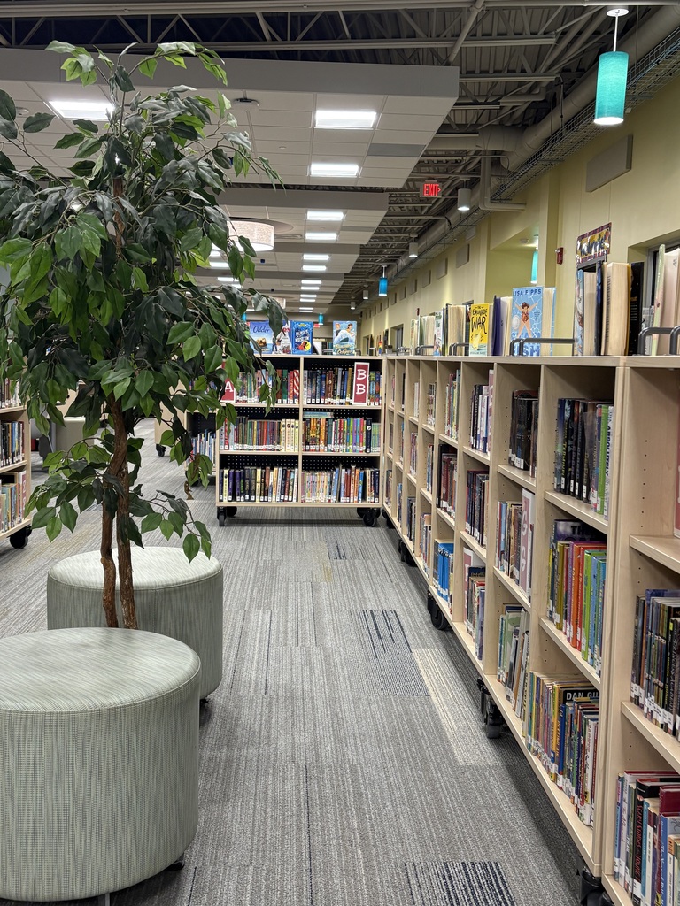Library interior with bookshelves filled with books, a tall plant, and round seating in the middle.