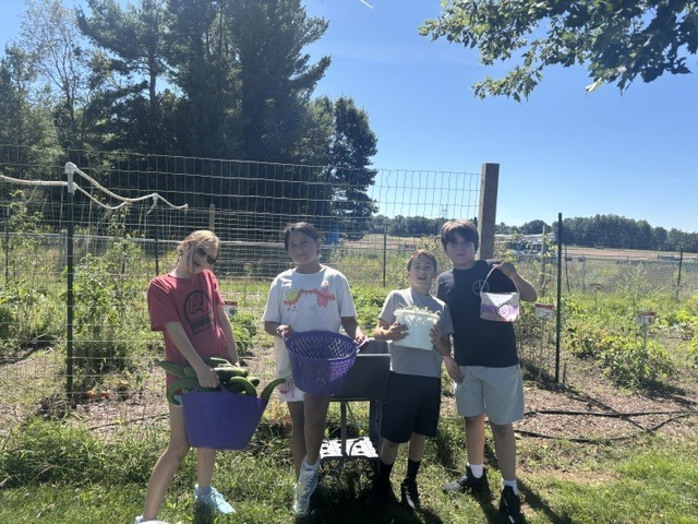 Four individuals stand in a garden. They hold baskets and a wheelbarrow. A fence and trees are in the background.