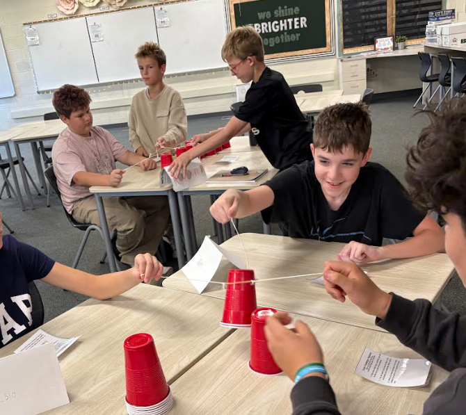 Students using a rubber band with three strings attached to move red solo sups