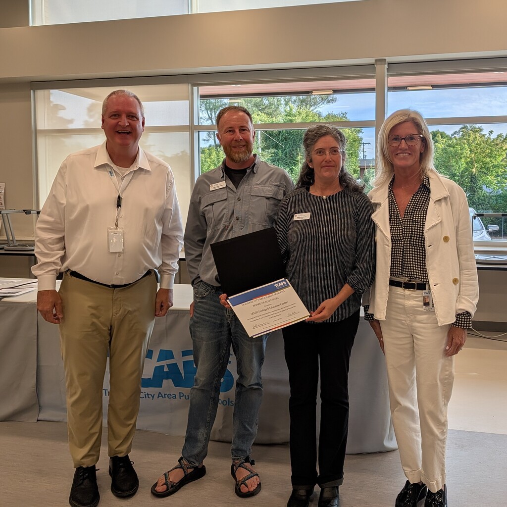 Four people stand together in a room, three women and a man. One woman holds a framed certificate.