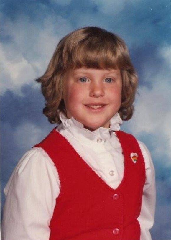 A child with short blond hair, wearing a red vest over a white shirt, smiles against a blue sky background.
