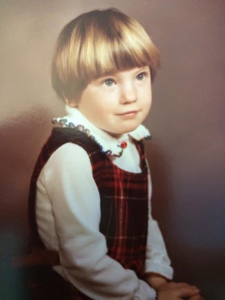 A child with short blonde hair wearing a white long-sleeve shirt and a red and black plaid vest.
