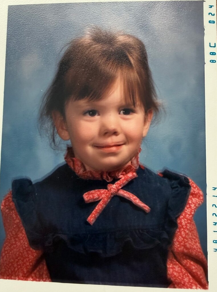 Young child with short hair, wearing a blue and red dress, smiling against a light blue background.