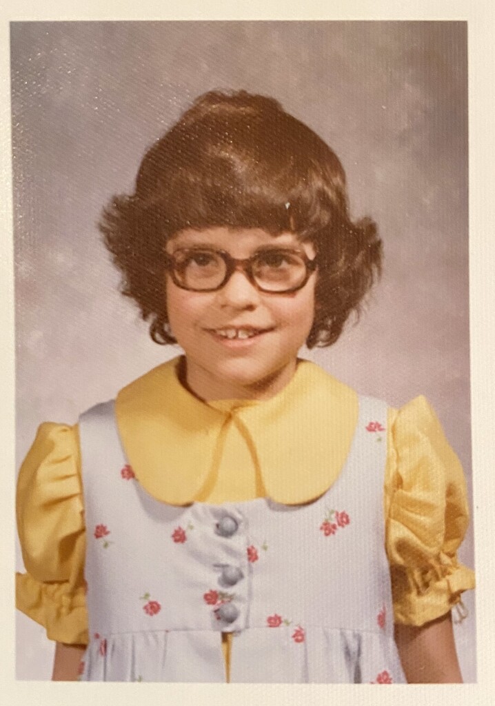 A young girl wearing glasses, a yellow collar, and a floral dress. She is smiling widely. Background is blurred.