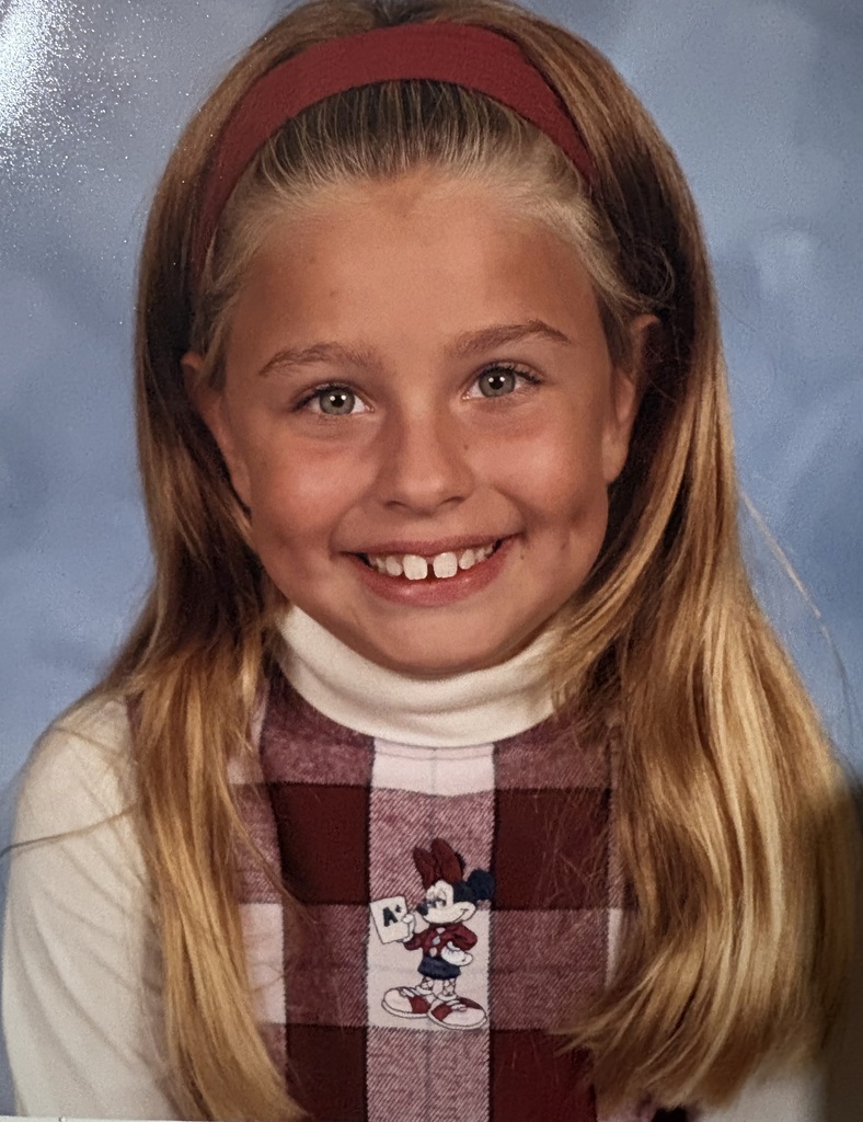A girl with long blonde hair, wearing a red headband and a checkered shirt, smiles for the camera.