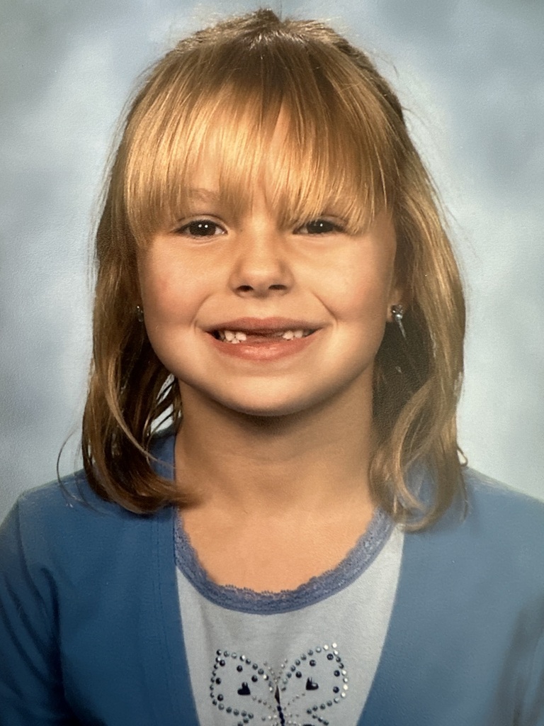 A young girl with blonde hair smiles brightly for a photo. She wears a blue top with a butterfly design.