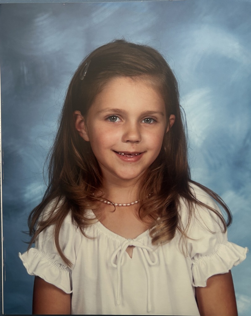 Girl with long hair smiles at camera. Wearing a white dress with ruffles. Background is blue.