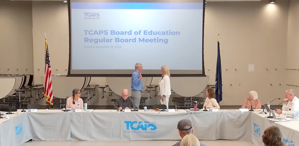 A conference room with a screen displaying "TCAPS Board of Education Regular Board Meeting". A group of people sit at a long table.