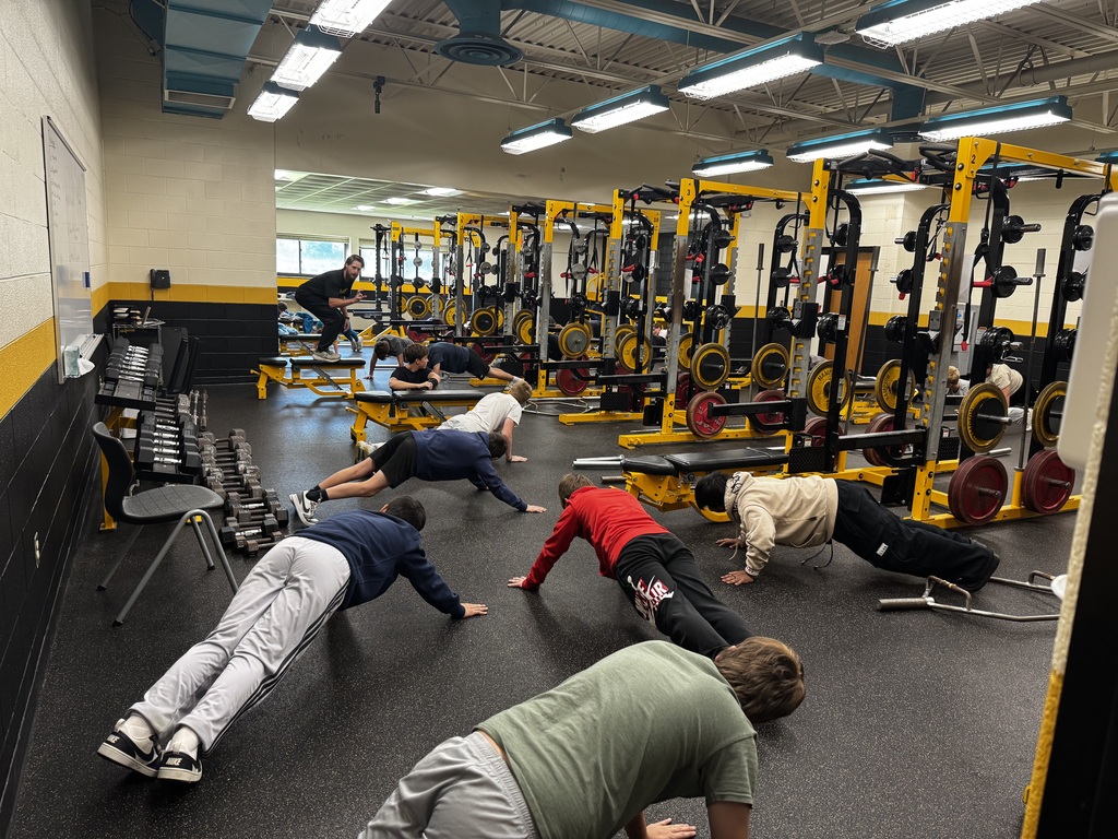 In a gym, several people are doing push-ups on the floor. Some are using equipment behind them.