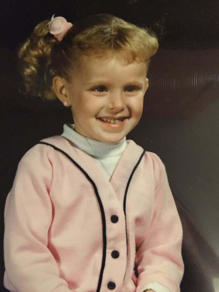A smiling young girl with pigtails and a pink jacket with black trim, seated against a dark background.