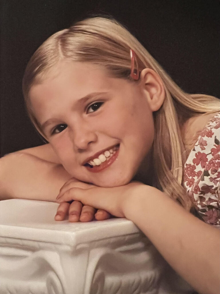 A young girl with blonde hair smiles, leaning on a white pedestal with floral-patterned clothing.