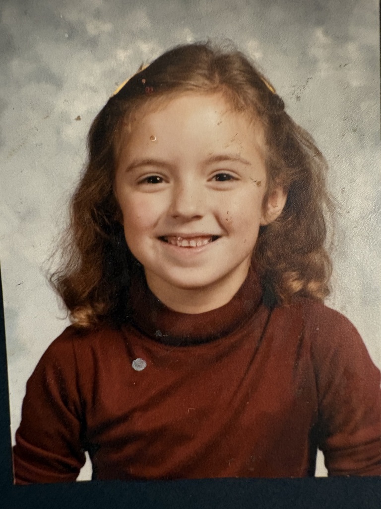 A young girl with brown hair smiles for the camera. She wears a brown turtleneck top and sits against a plain background.