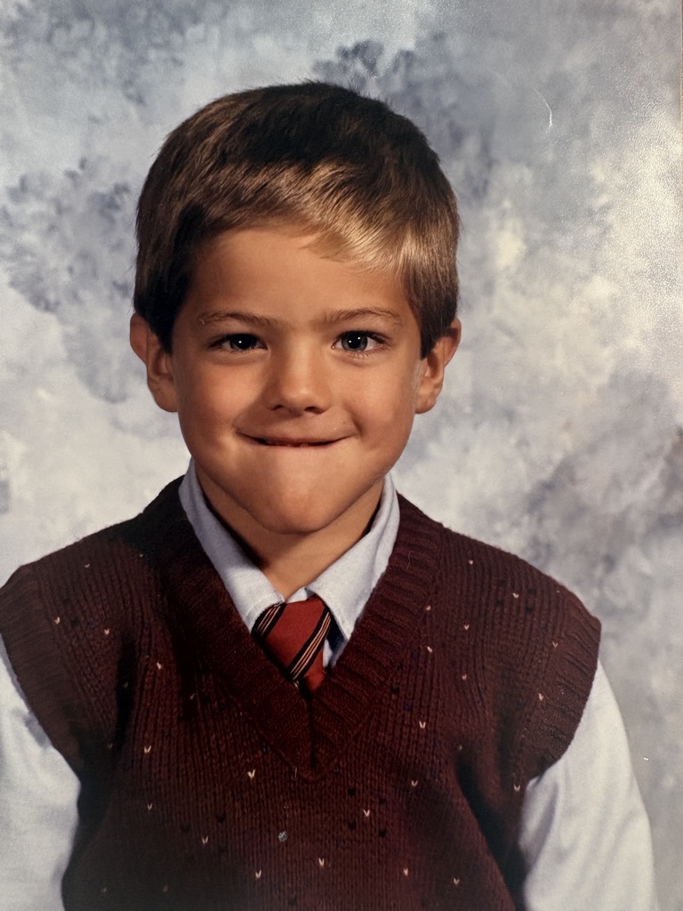 A young boy wearing a white collared shirt and a brown vest with a red and white tie.