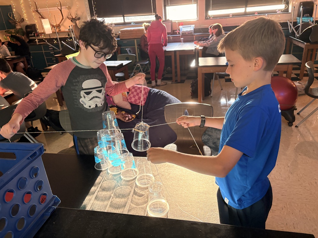 Two young boys in a classroom conduct an experiment with cups connected by string.