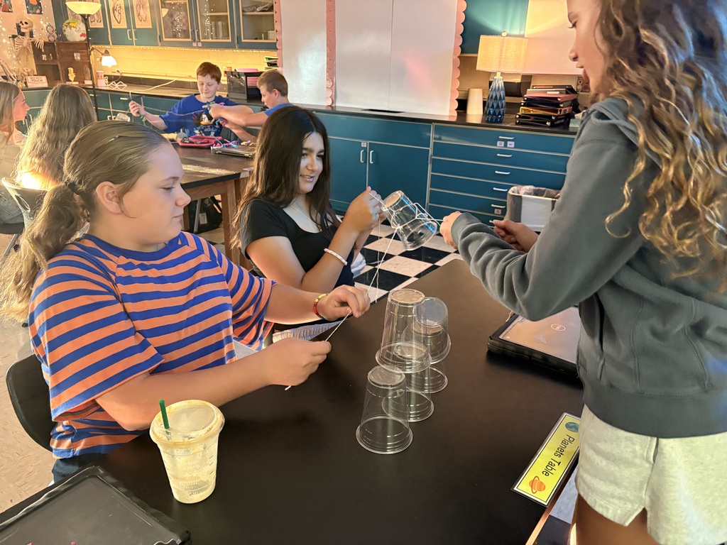 Two girls working on a science project at a table. One is pouring water into cups.