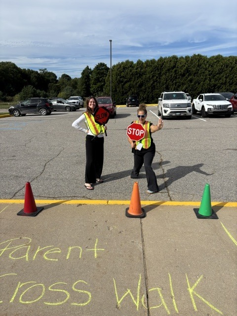 Two people in reflective vests stand in a parking lot, one holding a stop sign, with traffic cones and "Parent Cross Walk" written on the ground.