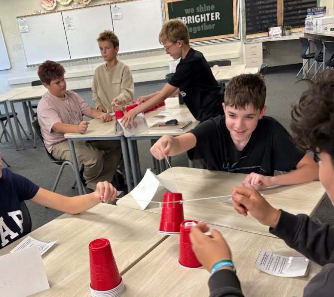 Students in a classroom setting, seated around tables, engaged in a group activity with red cups.