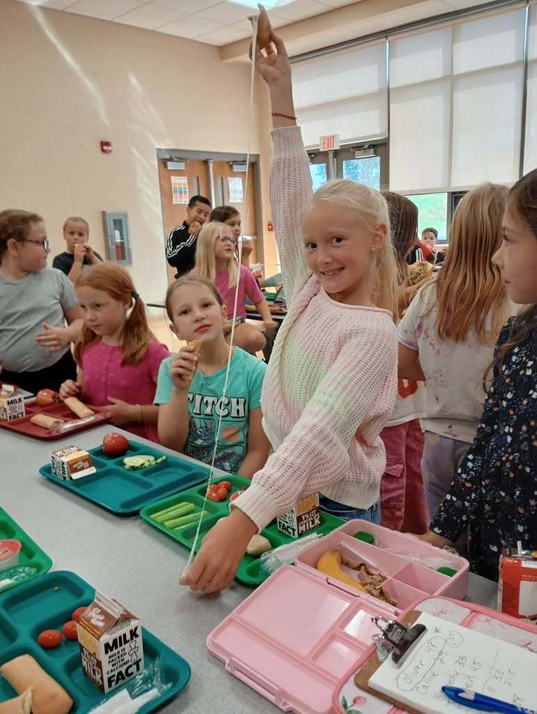Children sit at a cafeteria table. One child raises a hand holding a string with an attached apple.