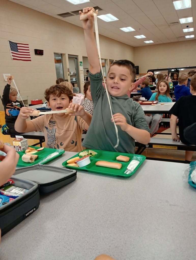 Children at a cafeteria table, with one child pulling a long piece of cheese. Trays of food and a flag in the background.