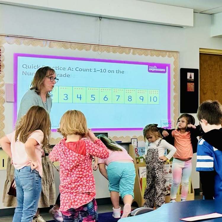 A classroom with a woman teaching a group of children in front of a projection screen showing numbers.