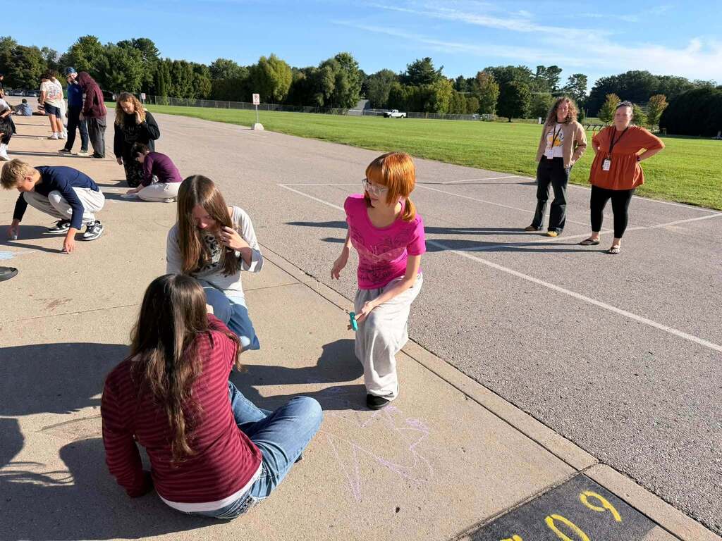 Several individuals are on a track, with a woman in a pink shirt walking across the track while others sit or crouch on the sidewalk.