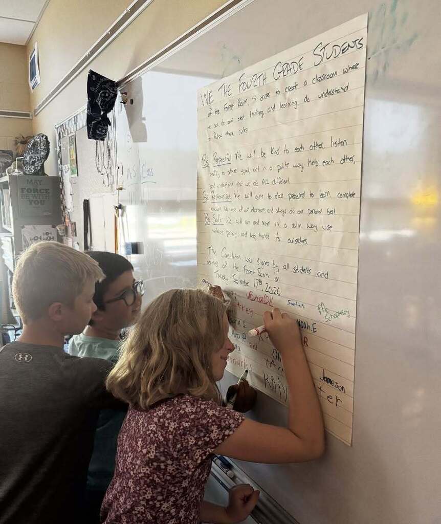 Three students in a classroom are looking at a whiteboard with writing. One student holds a marker.