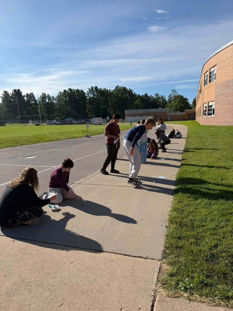 Several people skate, stand, and sit along a sidewalk with a grassy area and a building in the background.