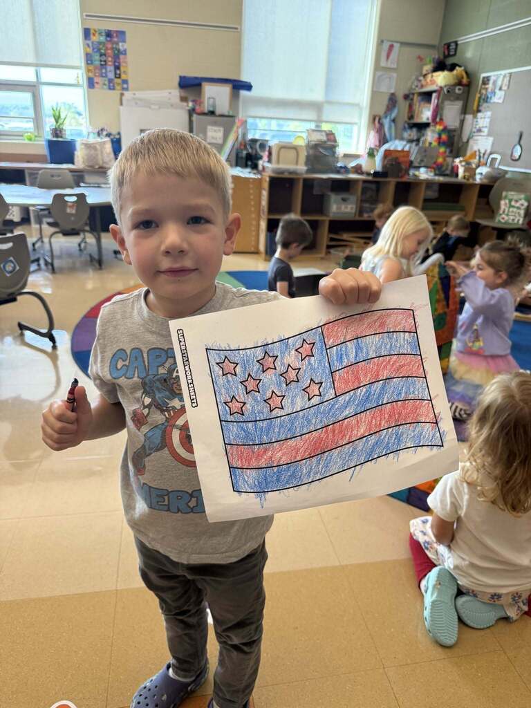 A young boy holding a paper flag with stars and stripes in a classroom with other children.