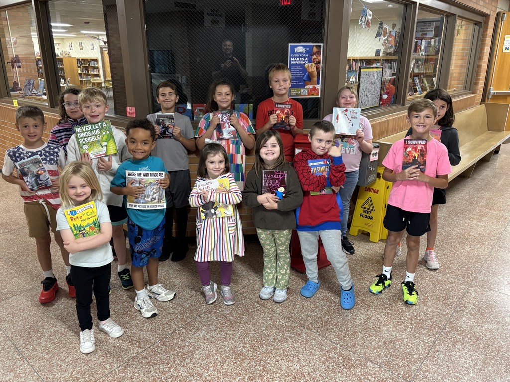 A group of children holding books poses for a photo in a school hallway.