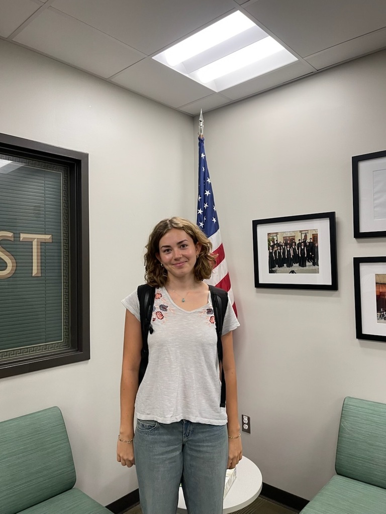 Woman in a white shirt and backpack stands in an office with a U.S. flag.