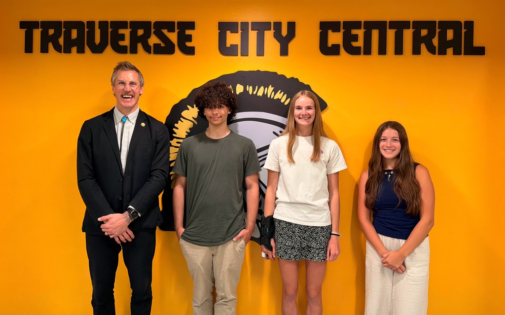 Four people stand in front of a yellow wall with the words "Traverse City Central" on it.