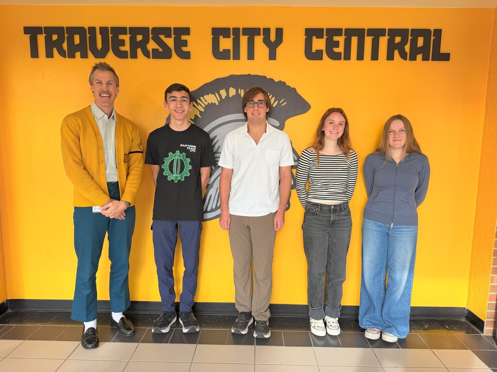 Five people pose for a photo in front of a yellow wall with the words "Traverse City Central".