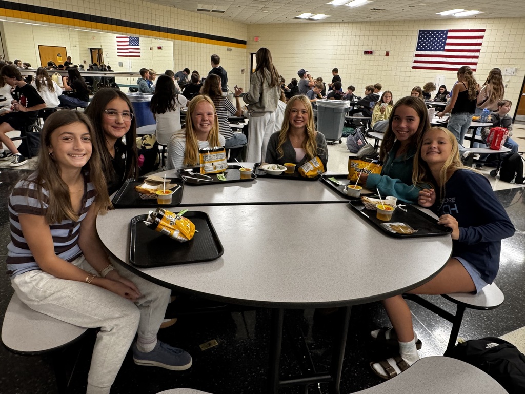 A group of girls sit around a cafeteria table with food, surrounded by a busy dining area with flags.