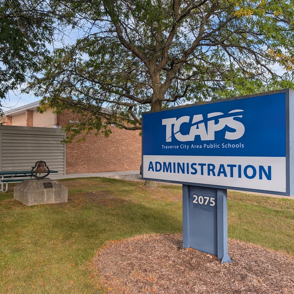 A sign reading "TCAPS Administration" is set on a grassy area. Behind it is a brick building with a bench.