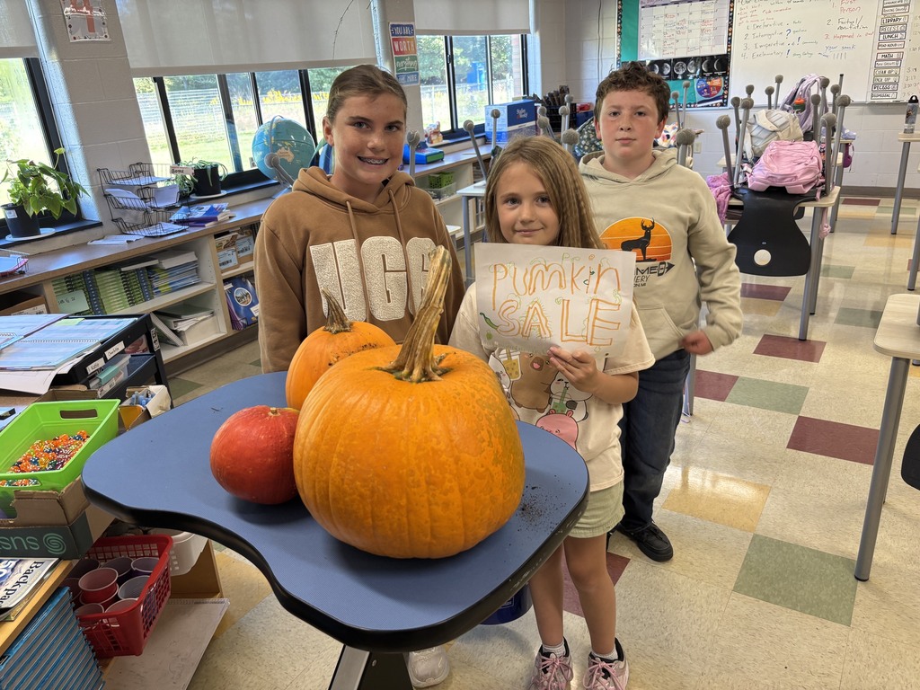 Three children in a classroom, one holding a large pumpkin, another with a smaller one, and the last holding a sign.
