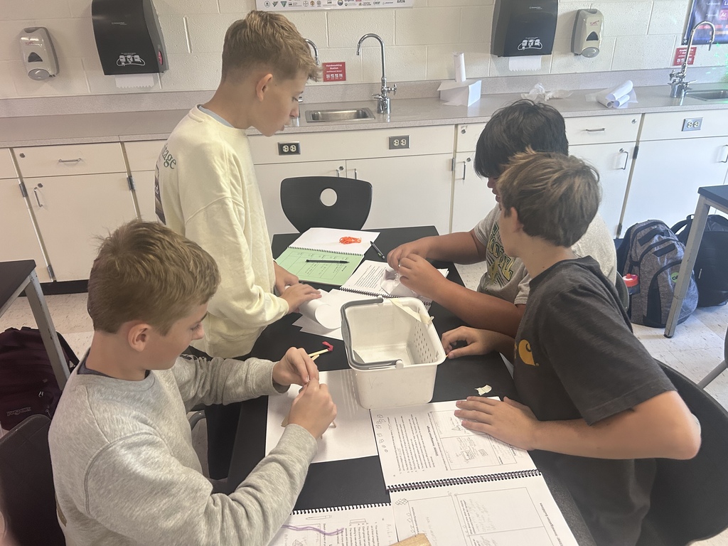 Four boys sit at a table, working on a project. A desk has papers, a container, and a marker.