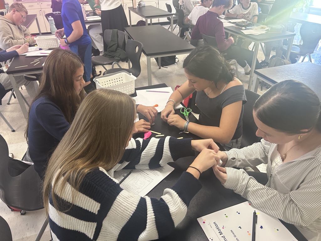 A group of students are seated around a table in a classroom, working on papers and scissors.